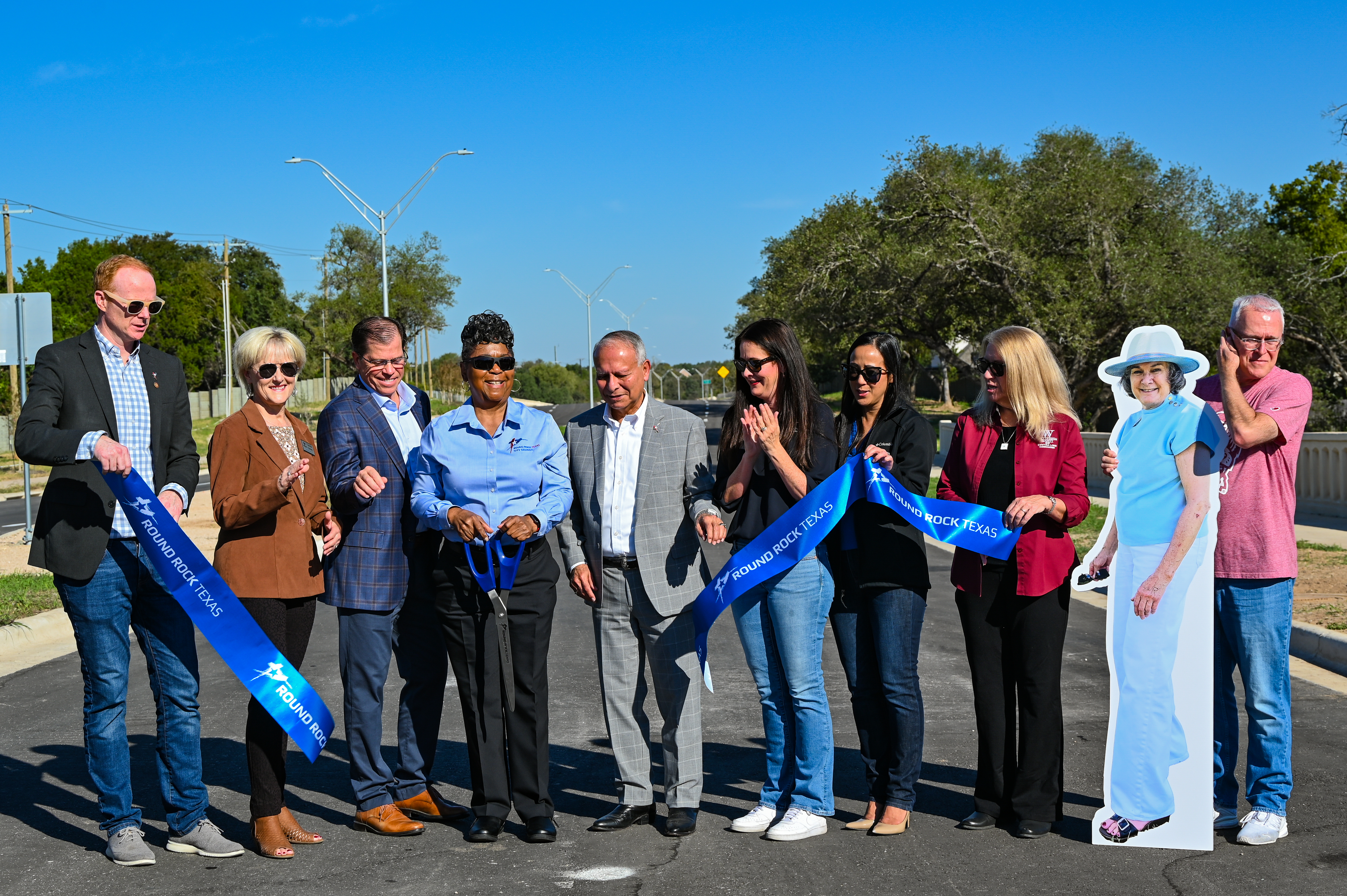 A group of people cutting a ribbon at an outdoor ceremony, with a life-sized cutout of a woman standing to the right.