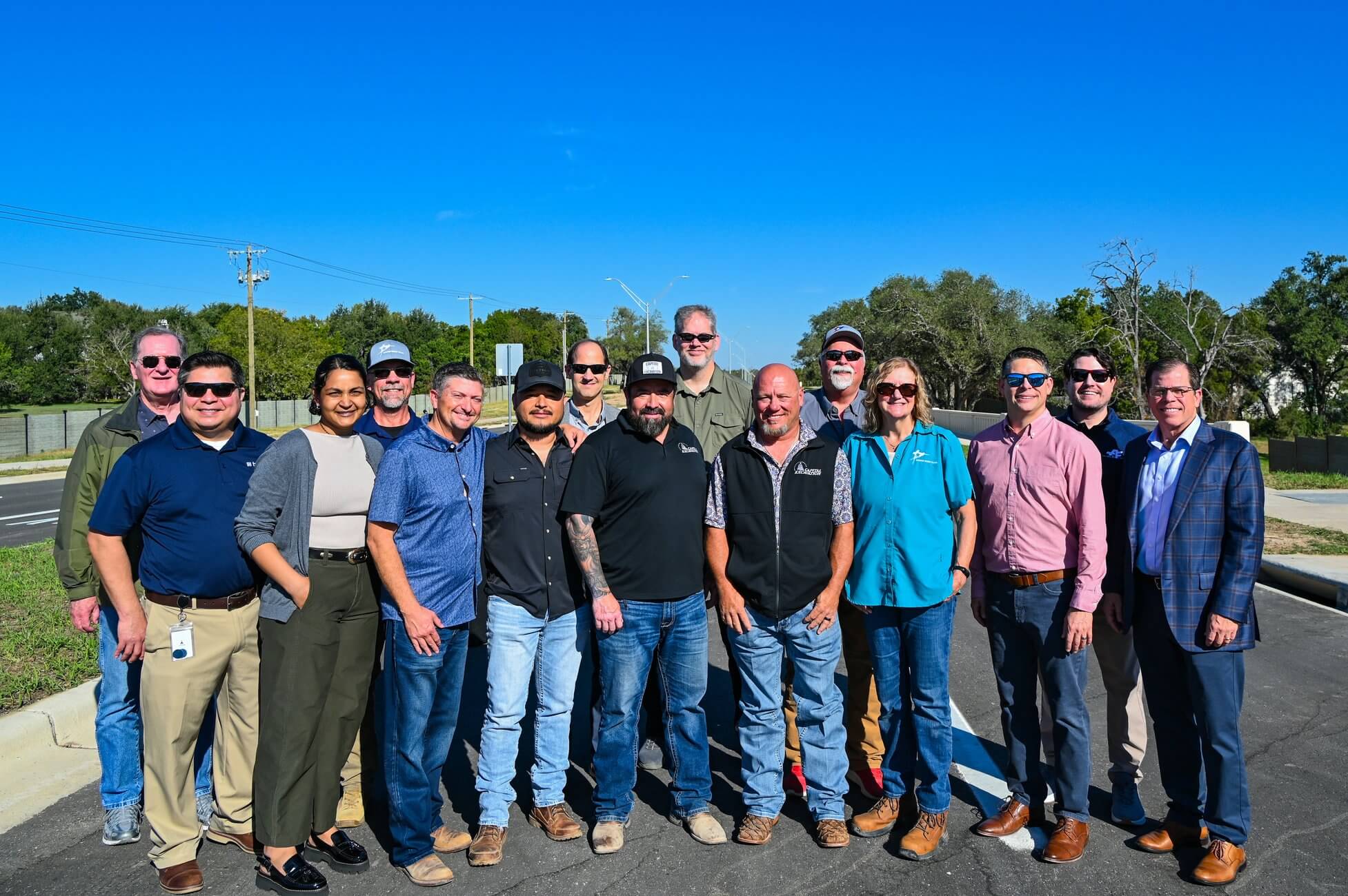 A group of thirteen people standing together outdoors on a sunny day.