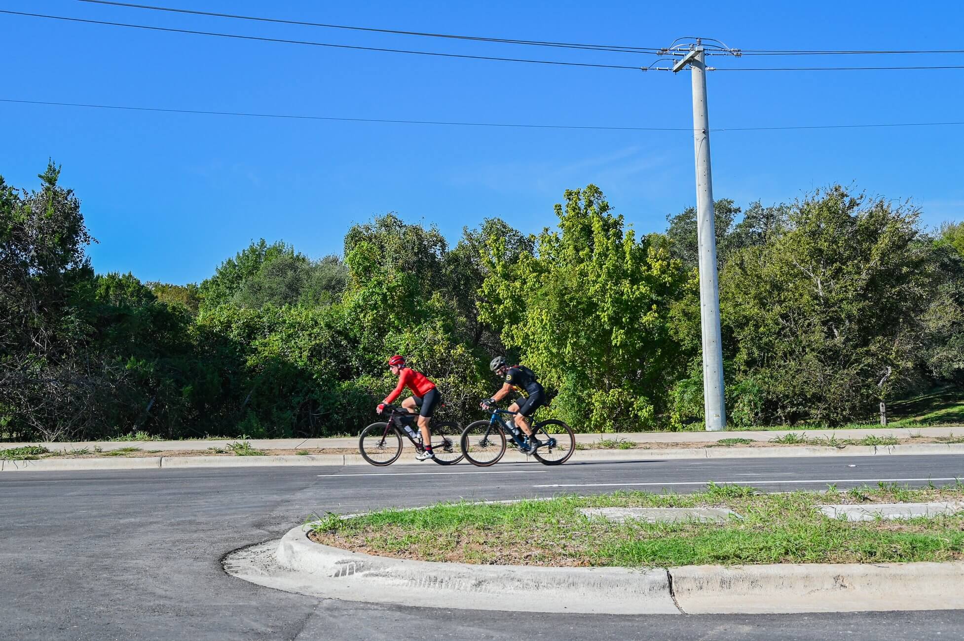 Two cyclists ride along a paved road next to a dense line of trees.
